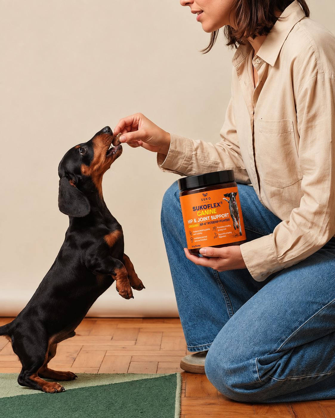 A woman kneels on a wood floor indoors, offering a treat to a black-and-tan dachshund while holding an orange and black container of SUKOFLEX® Dog Joint Support.